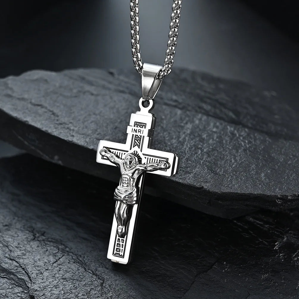 Colgante de cruz de Jesús en tono plateado con figura detallada, mostrado sobre una superficie de piedra oscura.