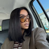 A woman with long black hair and sunglasses is seated inside a car, wearing a beige blazer.