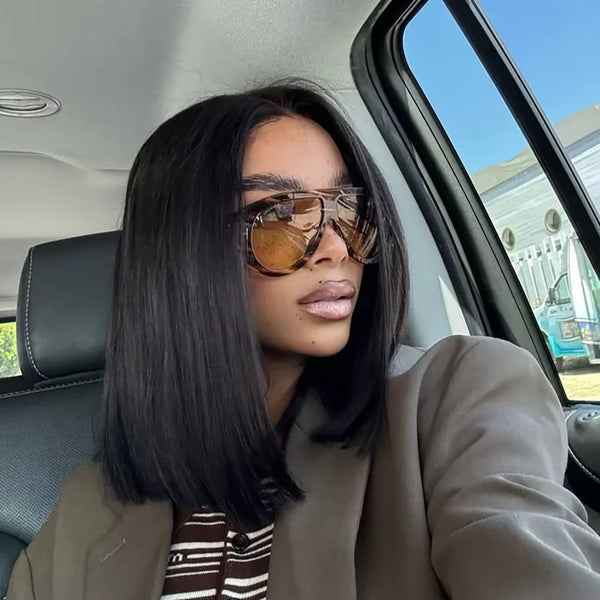 A woman with long black hair and sunglasses is seated inside a car, wearing a beige blazer.