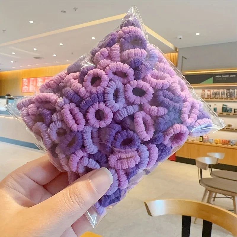 A hand holds a clear plastic bag filled with purple and lavender elastic hair bands for girls.