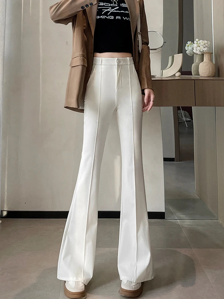 A woman models high-waist flare skinny white pants and a brown blazer in a modern interior.