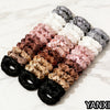 A set of five satin hair scrunchies in various shades of pink, beige, gray, and black arranged diagonally on a white marble s