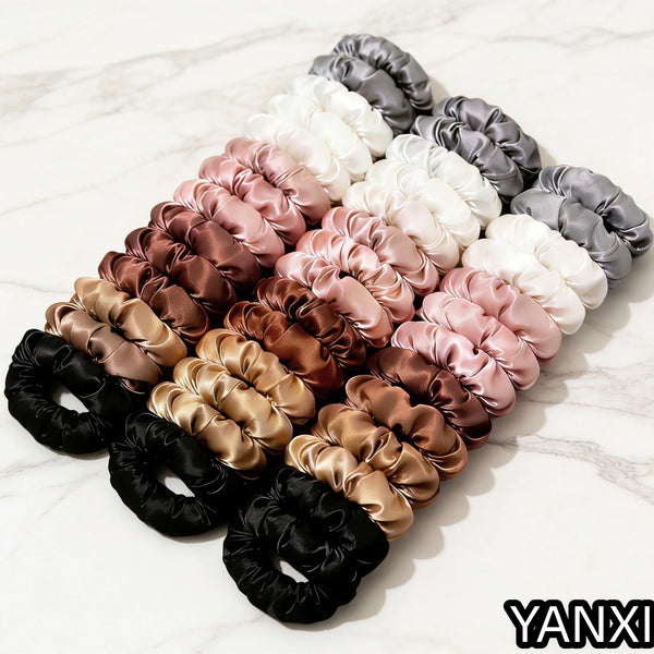 A set of five satin hair scrunchies in various shades of pink, beige, gray, and black arranged diagonally on a white marble s