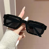 A hand holds a pair of black retro square fashion sunglasses against a light background.