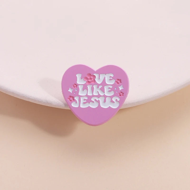 Broche émaillée en forme de cœur rose avec le texte Love Like Jesus et des fleurs roses et violettes.