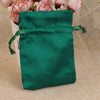 A dark emerald green satin drawstring pouch with a green tie, resting on a beige fabric next to pink flowers.