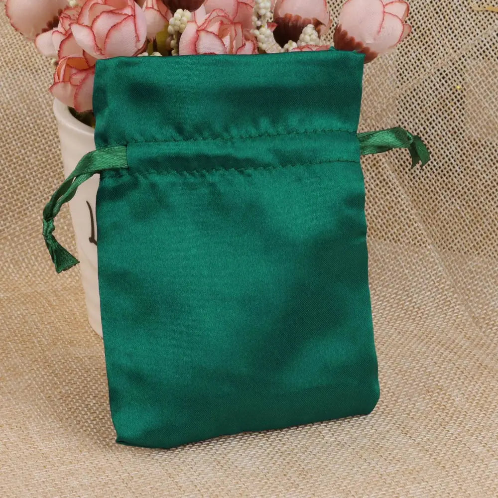 A dark emerald green satin drawstring pouch with a green tie, resting on a beige fabric next to pink flowers.