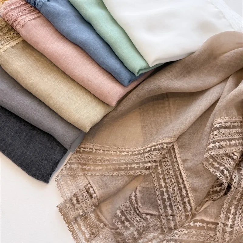 A stack of various colored fabric scarves and a beige bohemian lace trim shawl draped on a white surface.