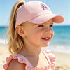 A smiling blonde girl wearing a pink unicorn baseball cap outdoors near the ocean.