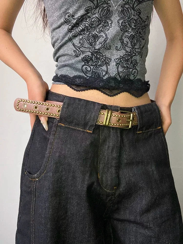 A close-up of a woman wearing dark denim pants and a grey top, featuring a brown belt with bronze rivets.
