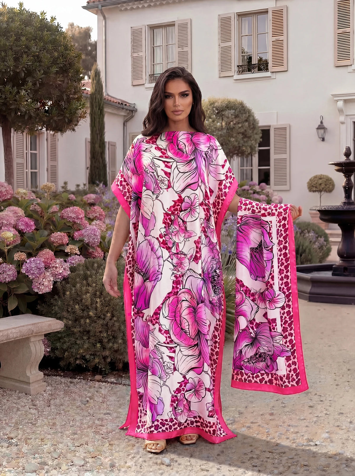 A woman models a pink and purple tropical floral print abaya scarf outdoors in a garden setting.