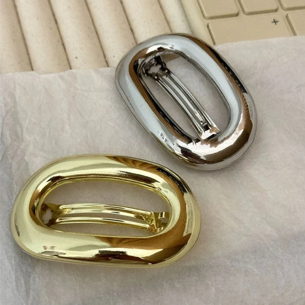 A pair of elegant metal oval hair clips, one silver and one gold, resting on white paper.