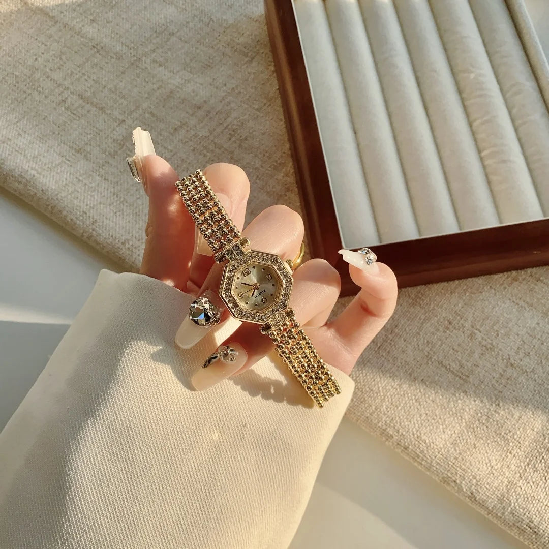A hand wearing an elegant quartz fashion wristwatch with a gold-toned band and diamond accents, set against a light backgroun