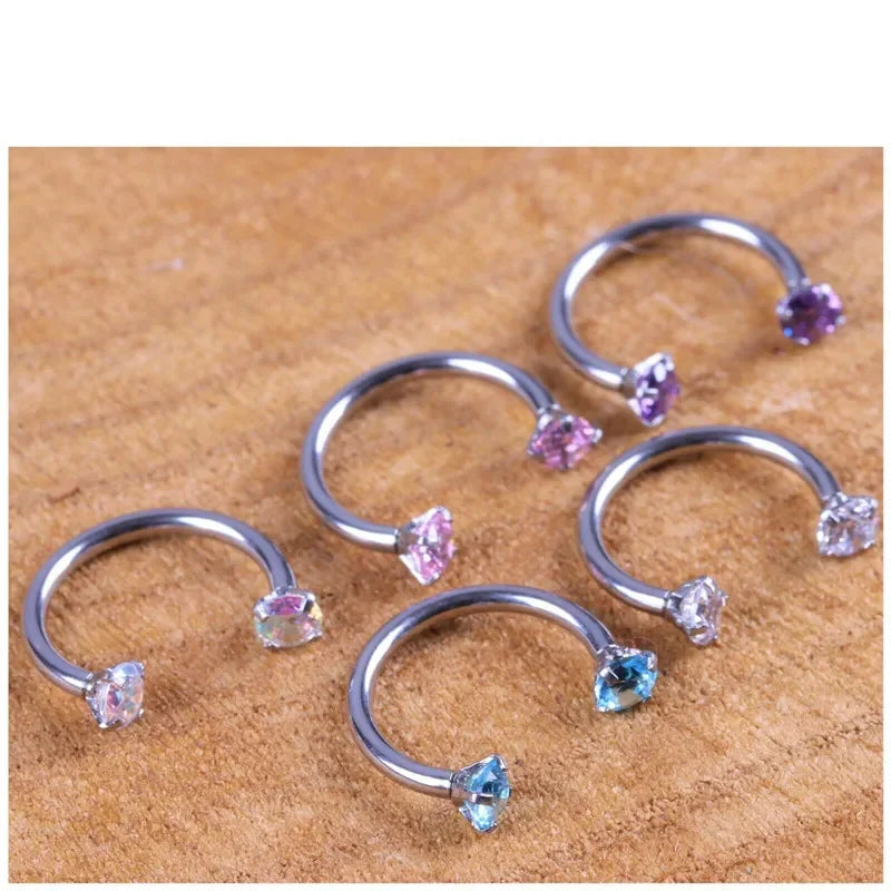 A collection of stainless steel crystal hoop ear piercings with various colored stones on a wooden surface.