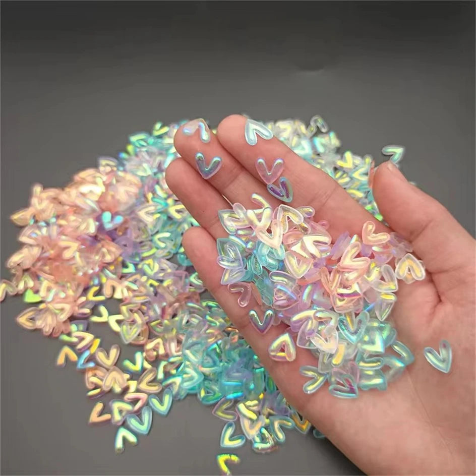 A hand holds a collection of iridescent, pastel resin nail art charms in various shades of pink, blue, and purple.