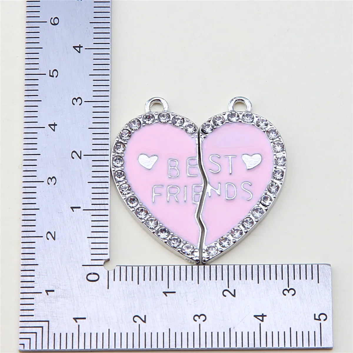 Colgante plateado en forma de corazón con esmalte rosa grabado con la palabra MEJORES AMIGOS, medido contra una regla.