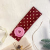 A pink silicone hairband with a small flower detail is displayed on a maroon fabric with gold dots.