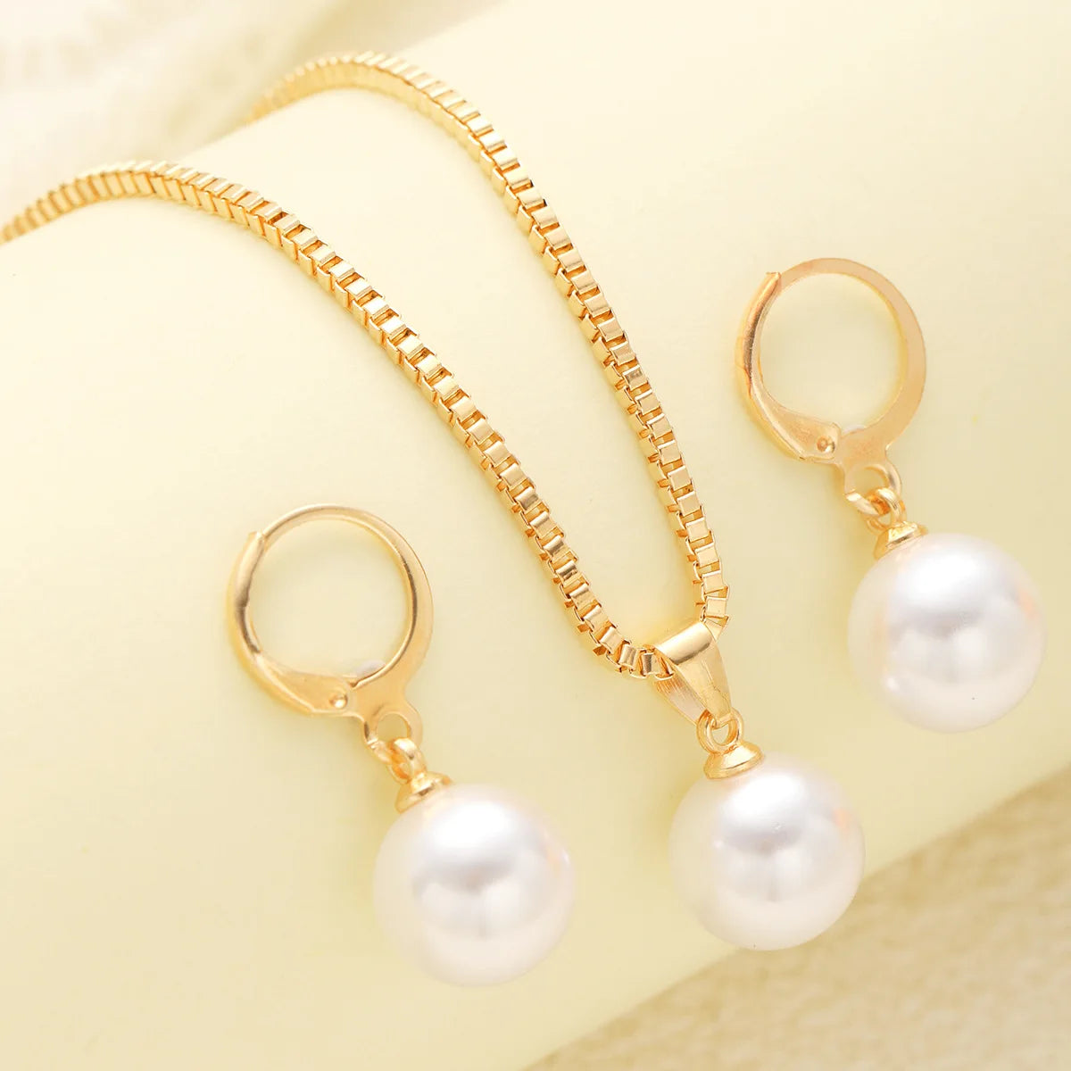 A set of dainty gold pearl jewelry featuring matching earrings and a delicate necklace displayed on a pale yellow background.