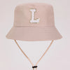 A light beige sun hat with a gold letter L embroidered on the front and a light tan strap.