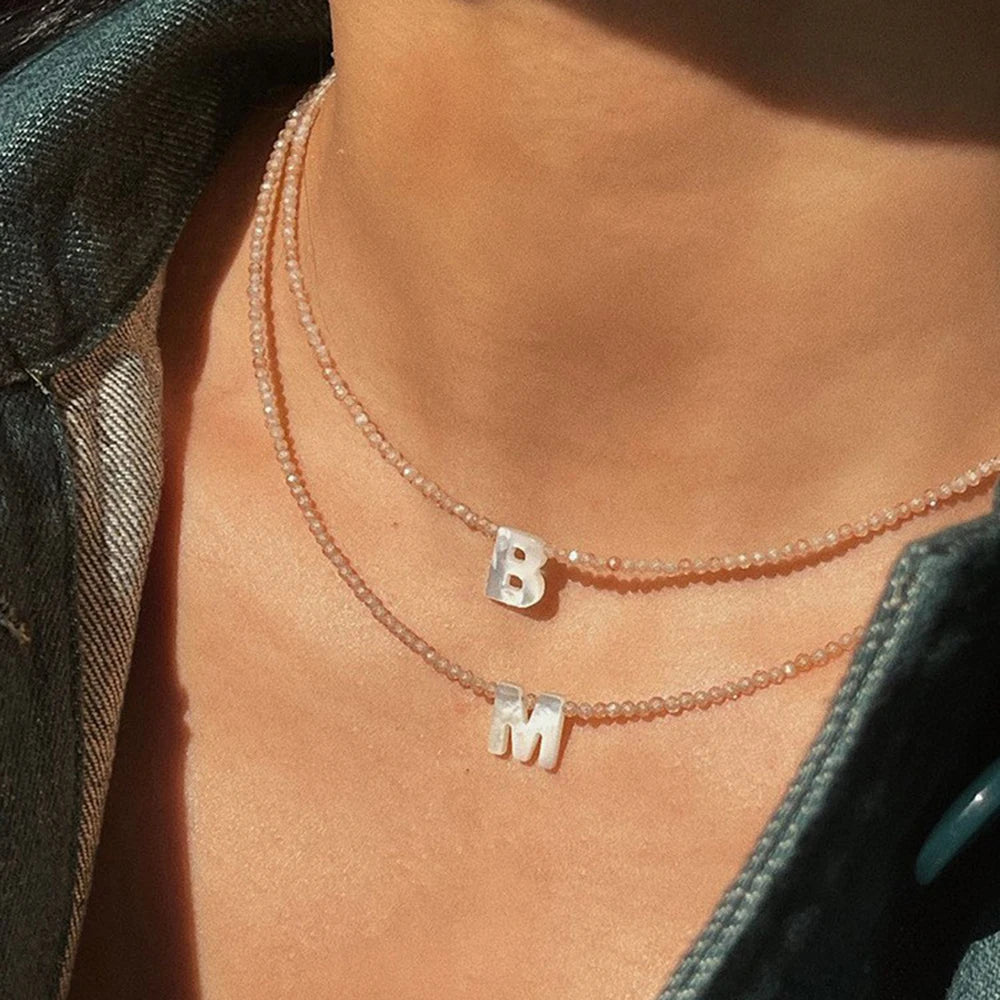A delicate necklace featuring a pearl chain with initial charms B and M worn against bare skin.