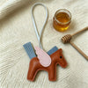 A small, handcrafted leather horse charm with pink and blue accents rests on a cream-colored knitted fabric next to a glass j