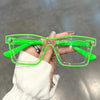A hand holds a pair of bright green, square-framed costume glasses with clear lenses.