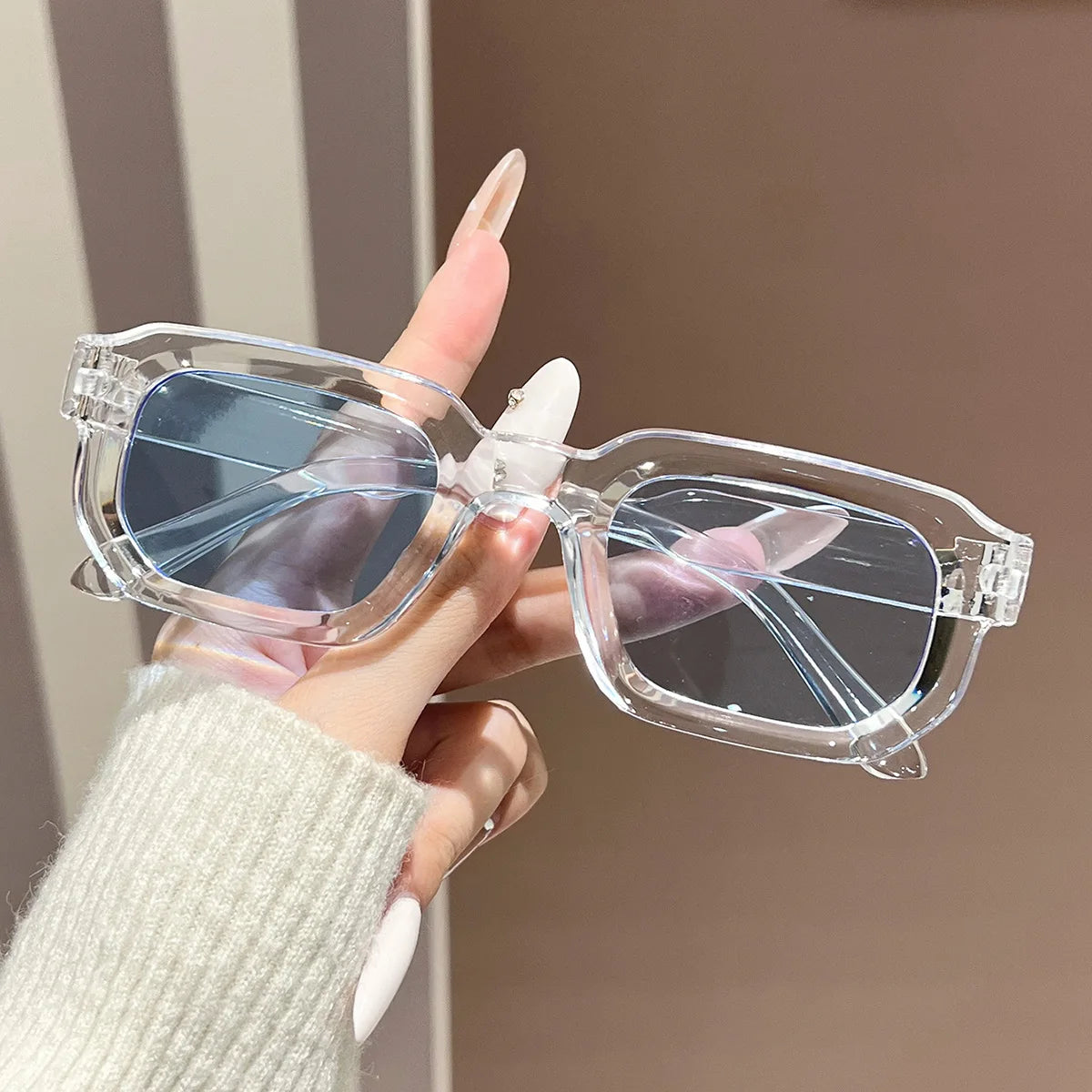A hand holds a pair of clear plastic retro square fashion sunglasses with blue tinted lenses.