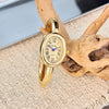 A gold-toned quartz watch with a round face and Roman numerals is displayed on a light fabric next to a piece of driftwood.