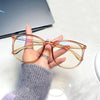 A hand holds a pair of large square anti-blue light spectacles with translucent orange frames.