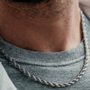 Gros plan du cou d'un homme portant un collier de corde tressée en acier inoxydable plaqué or 18K sur un t-shirt gris.