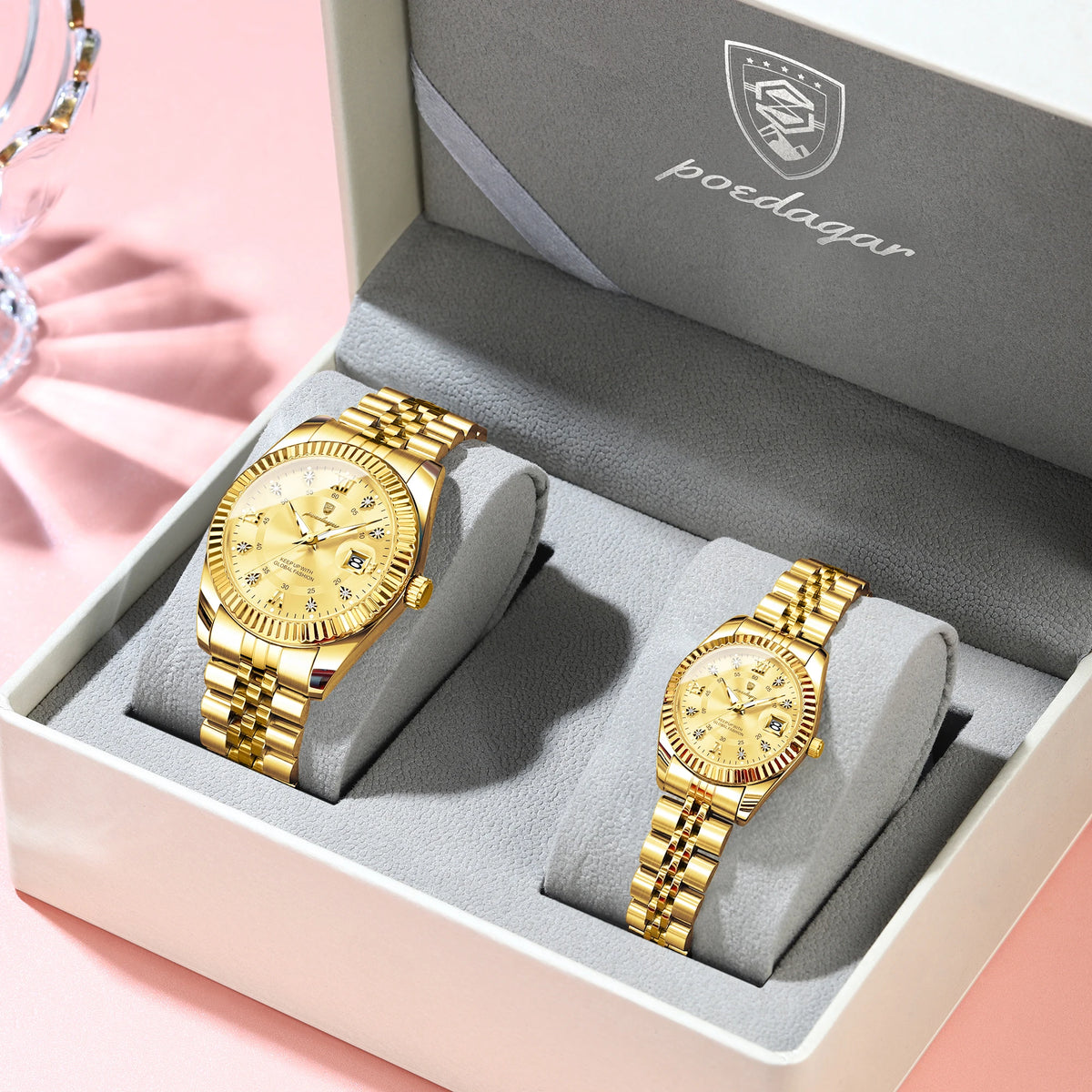 A pair of matching gold-toned watches with luminous hands and date display are displayed in a light gray box.