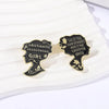 A pair of black enamel lapel pins shaped like a girl's head with text reading OBSTINATE HEADSTRONG GIRL and SHE IS TOO fond o