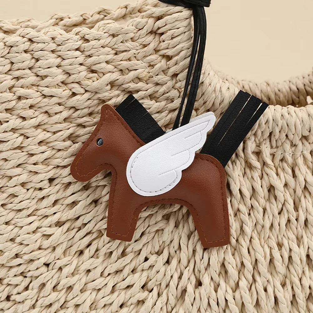 A brown horse pendant charm with white wings attached to a black strap, resting on a cream-colored knitted fabric.