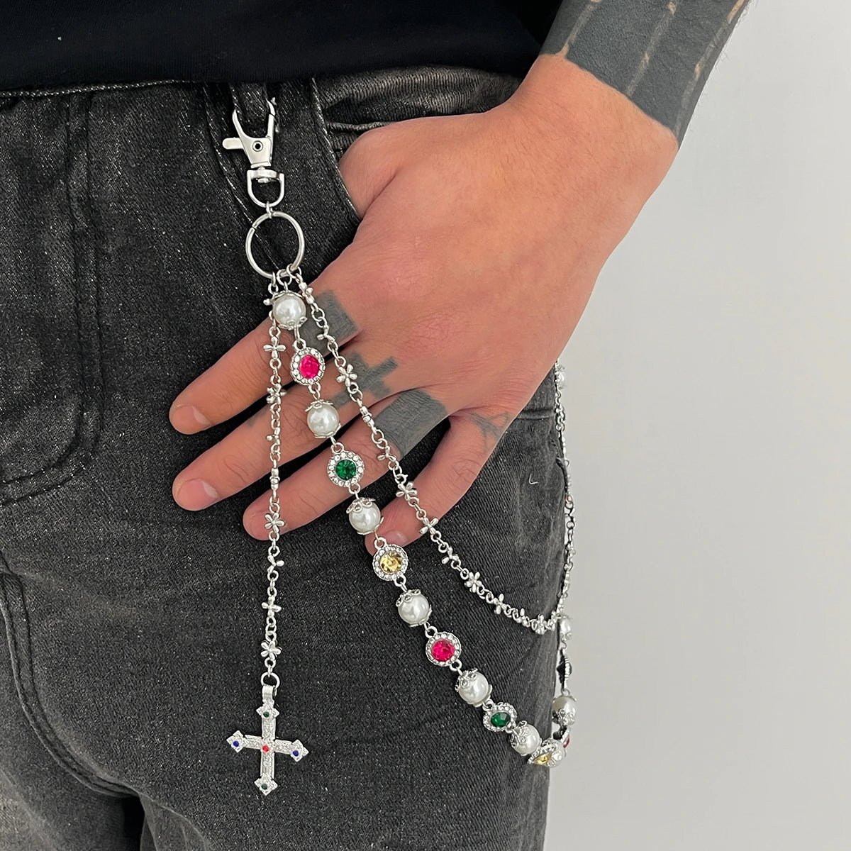 A hand holds a silver pearl cross pendant keychain against dark denim fabric.