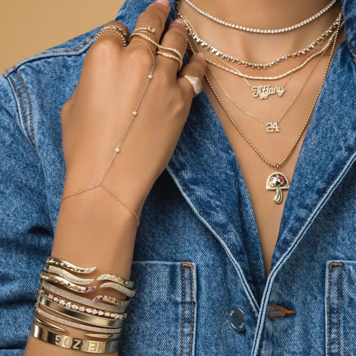 A close-up of a hand wearing multiple layered gold and silver necklaces and a geometric bezel charm bracelet against a denim 