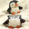 Un pingüino de peluche con pelaje gris y blanco, sentado sobre una superficie crema, protegiendo unos auriculares blancos den