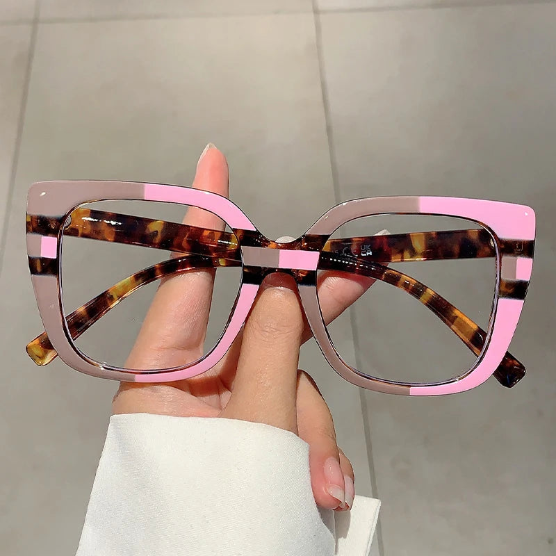 Eine Hand hält eine modische, große, quadratische Brille mit Schildpatt- und rosa Akzenten.