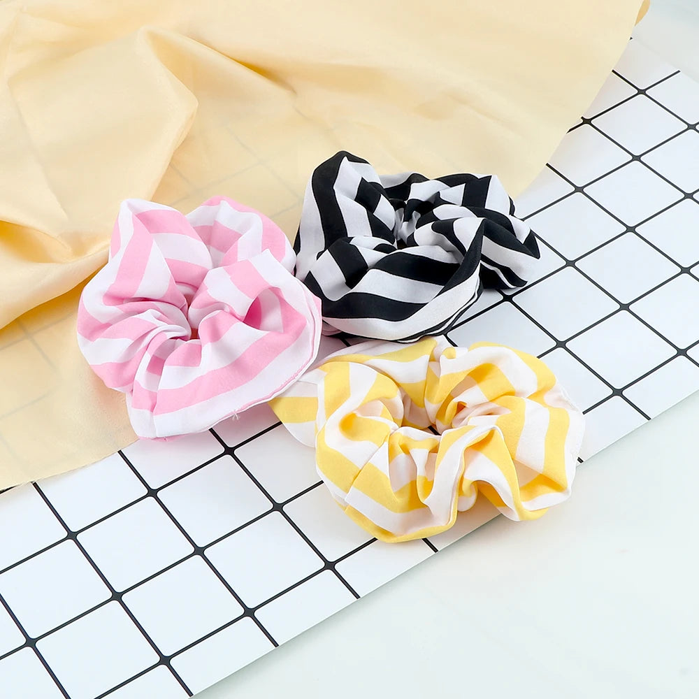 A collection of three colorful knitted hair scrunchie headbands in pink, black and white stripes, laid on a grid background.