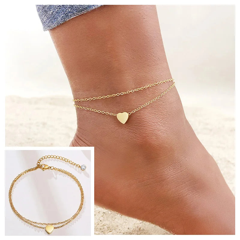Gold stainless steel snake chain anklet with a heart charm worn on a woman's ankle.