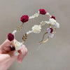 A hand holds an elegant rose vine headband hair accessory featuring deep red and cream roses and gold accents.