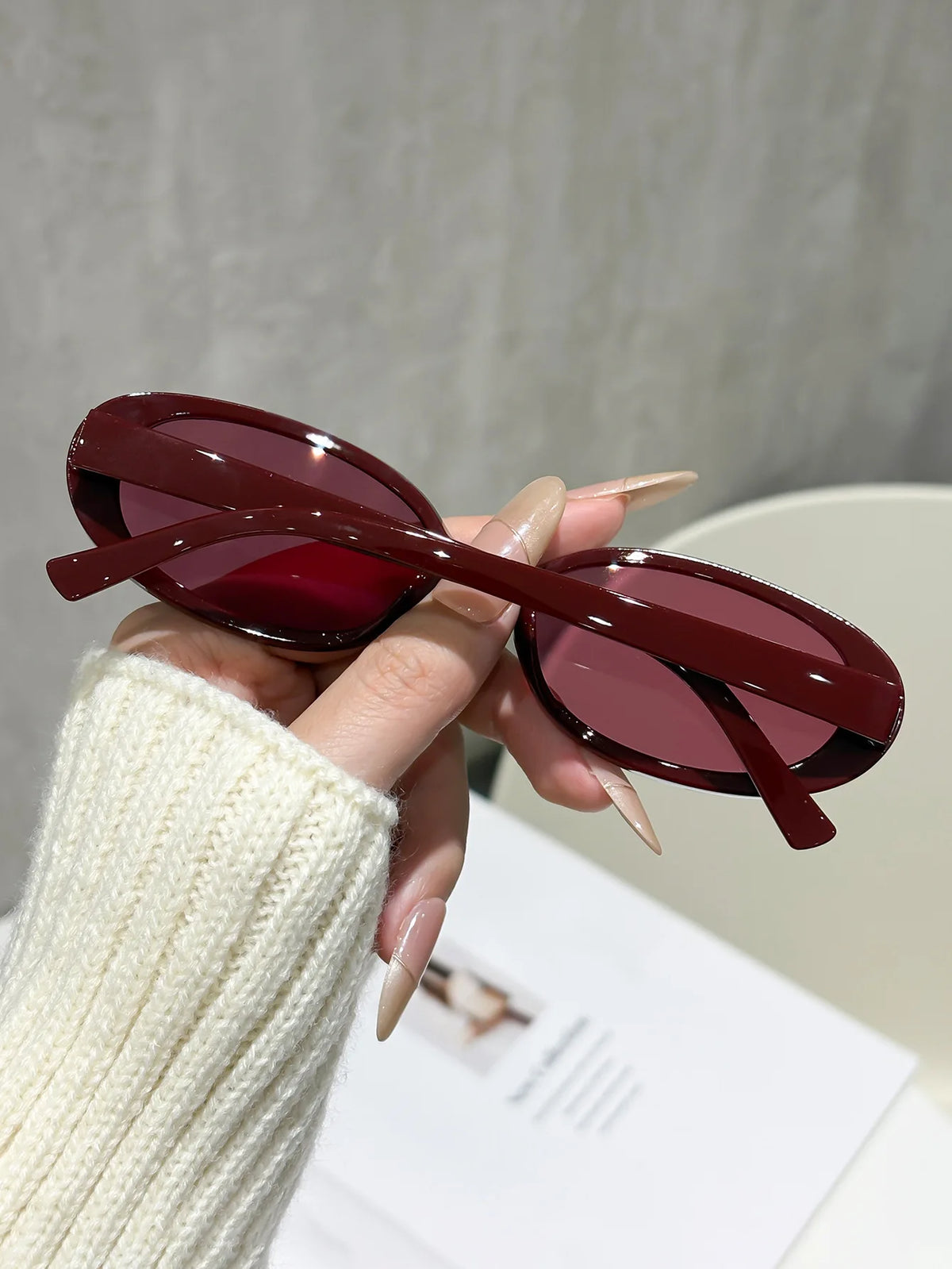 A hand holds a pair of maroon cat eye sunglasses with thin brown frames against a light background.