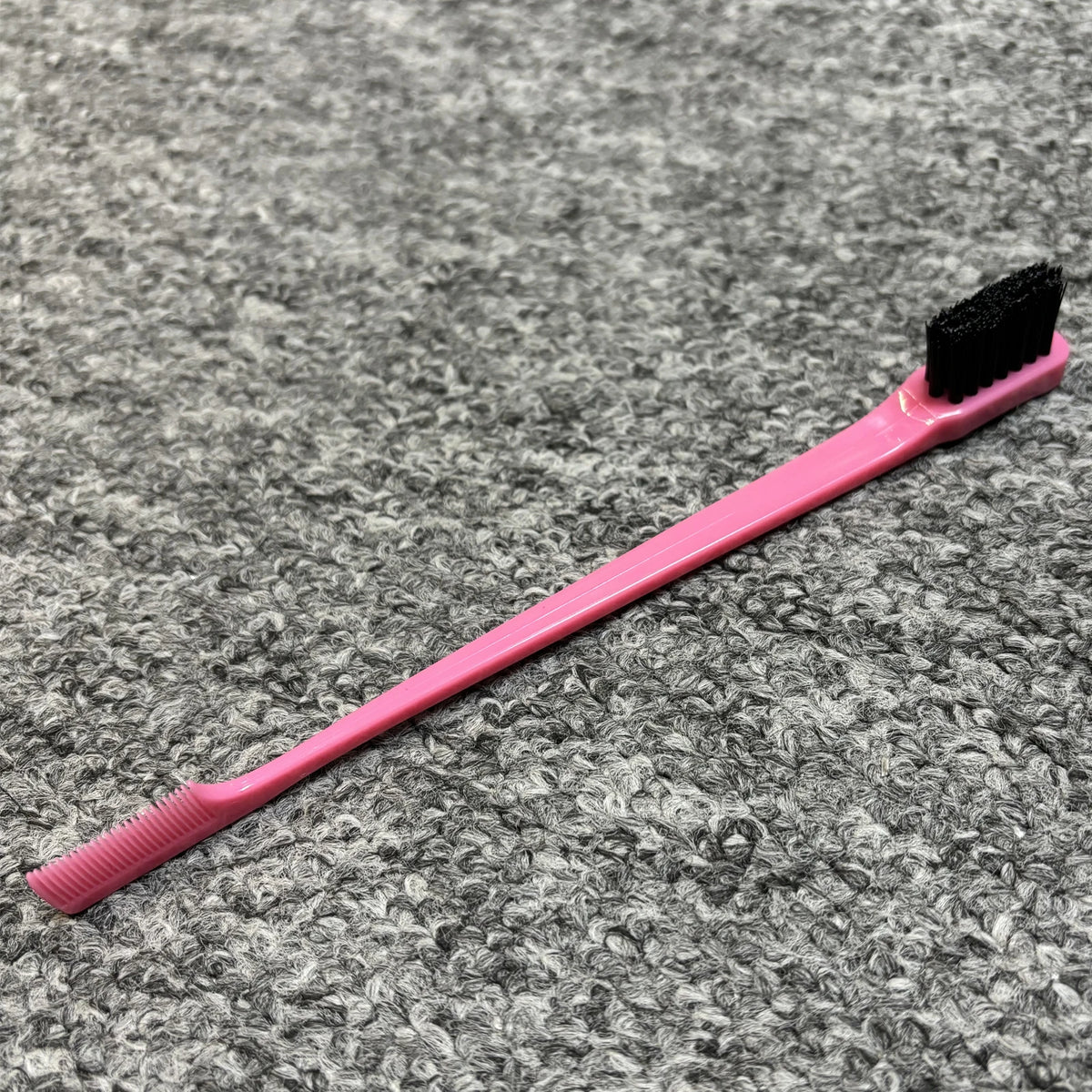 Pinkes professionelles, doppelseitiges Augenbrauenkamm mit schwarter Bürste auf grauer strukturierter Oberfläche.