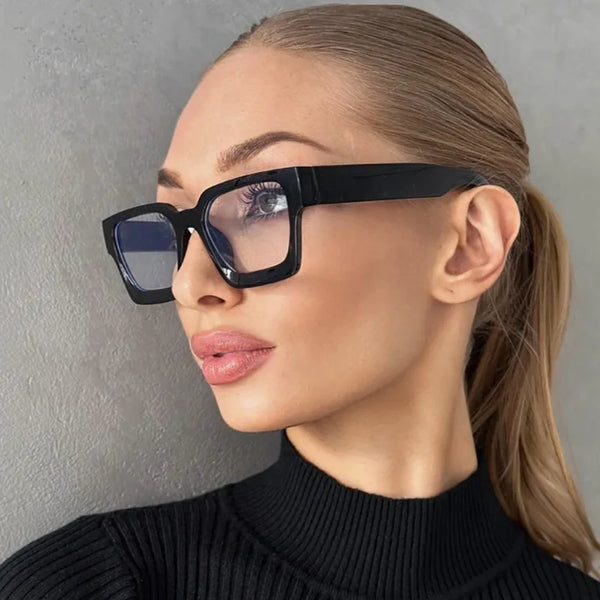 A woman wearing black vintage square eyeglasses against a gray background.