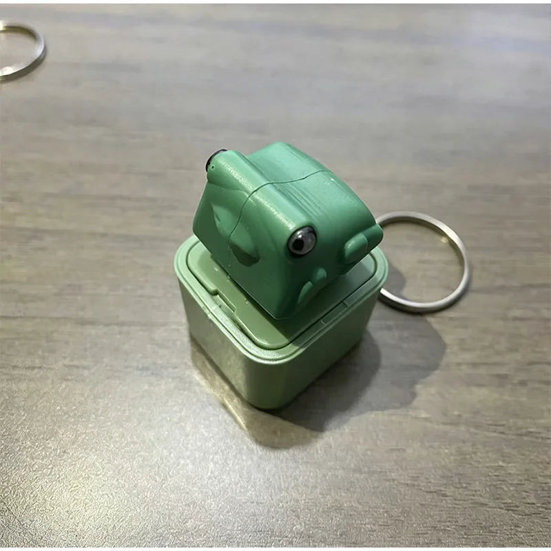 A light green fidget frog keychain with a clear plastic case resting on a wooden surface.