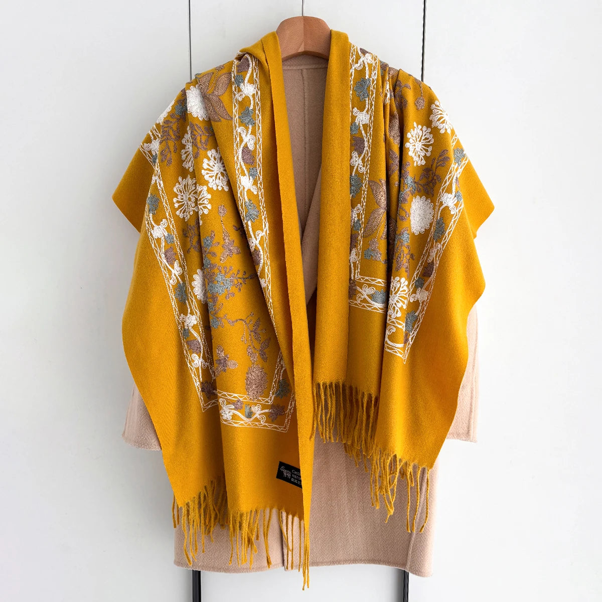 A mustard yellow luxury floral scarf with beige trim and fringe draped over a hanger.
