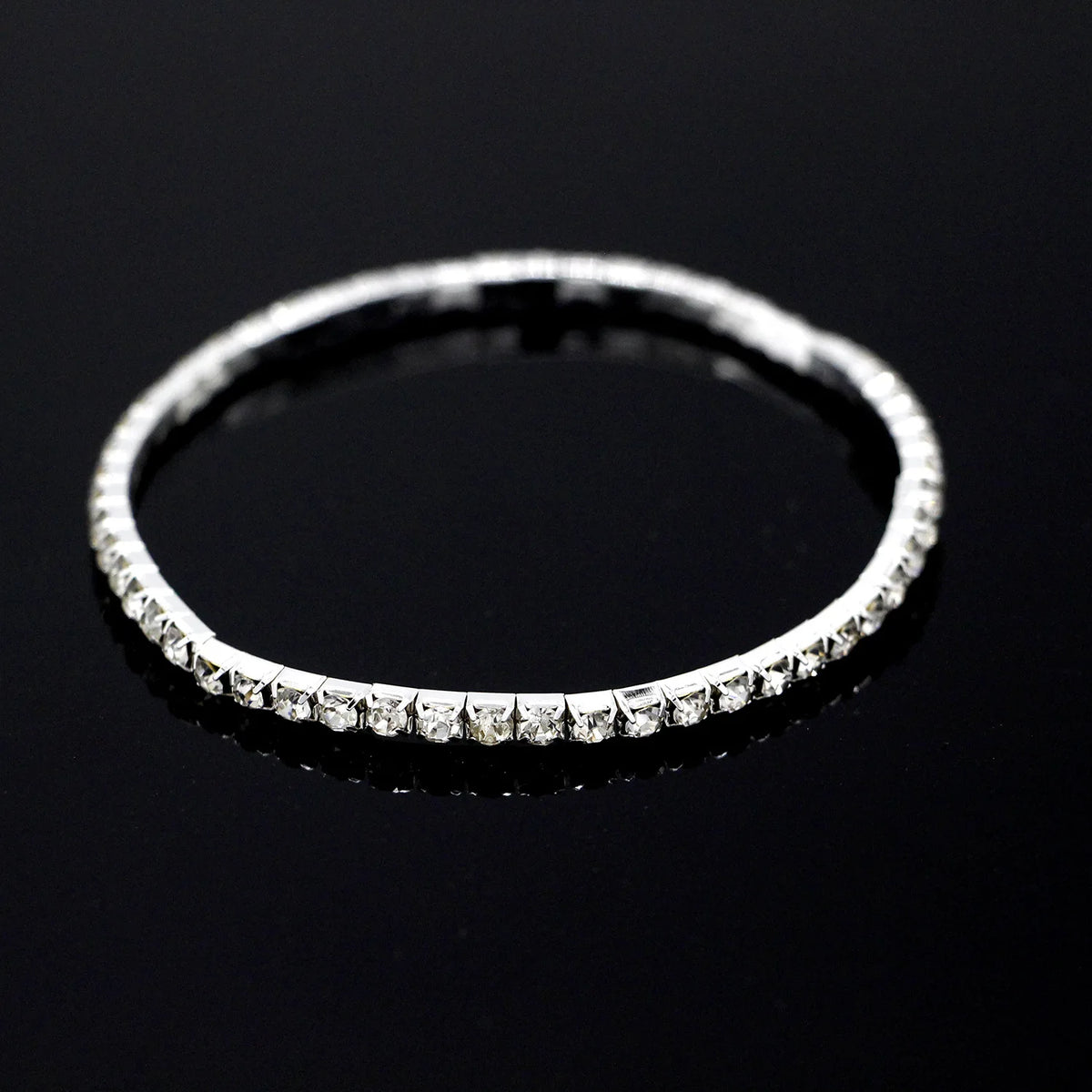 A close-up shot of a silver tennis anklet featuring small, clear crystals on a black reflective surface.