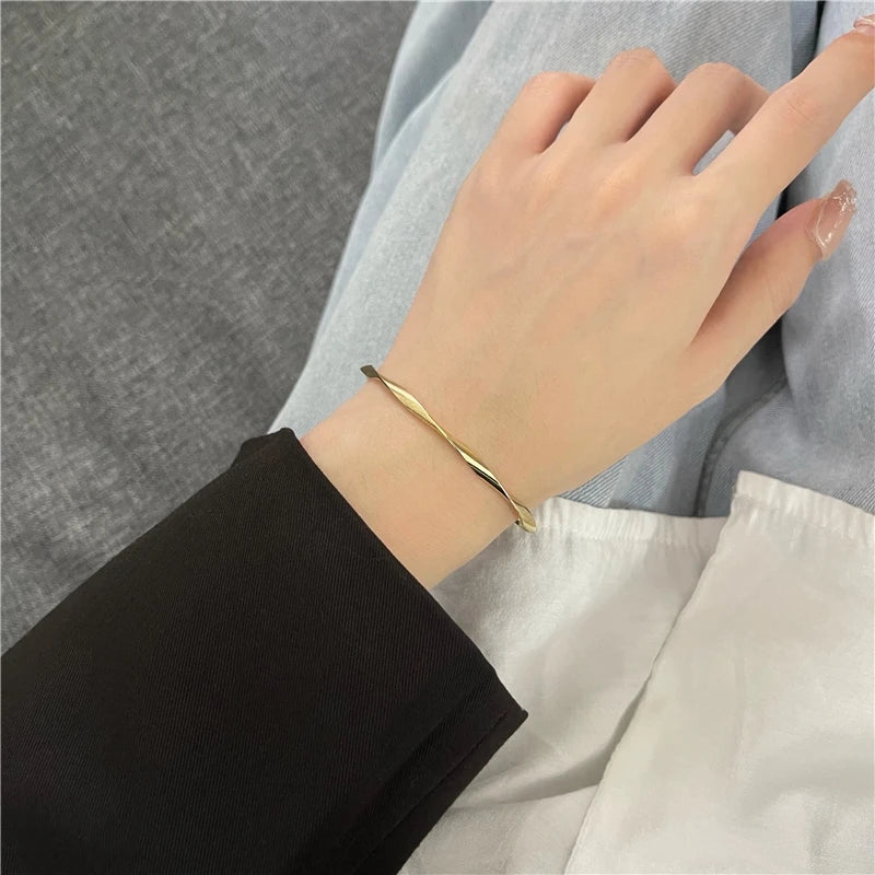 A hand wearing a minimalist gold-tone twisted cuff bangle, shown against a light blue fabric background.