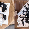 A black and white floral print silk scarf draped on a beige fabric next to a brown leather sofa.