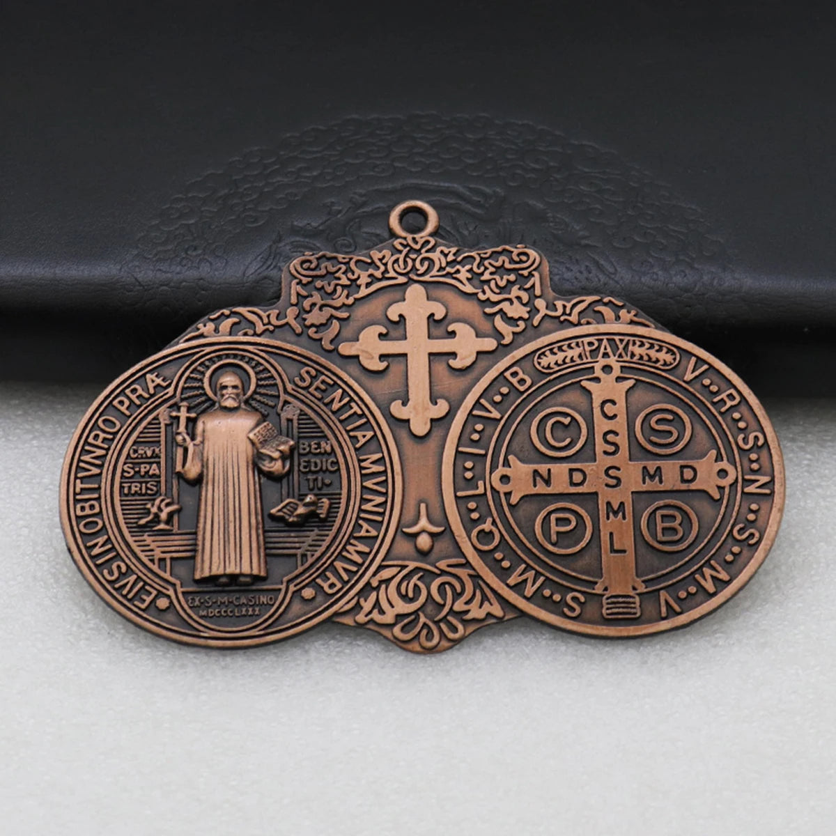 A bronze Saint Benedict protection medal featuring a cross and religious symbols, displayed on a dark surface.