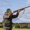 A person wearing a camouflage thermal beanie is aiming a rifle outdoors.